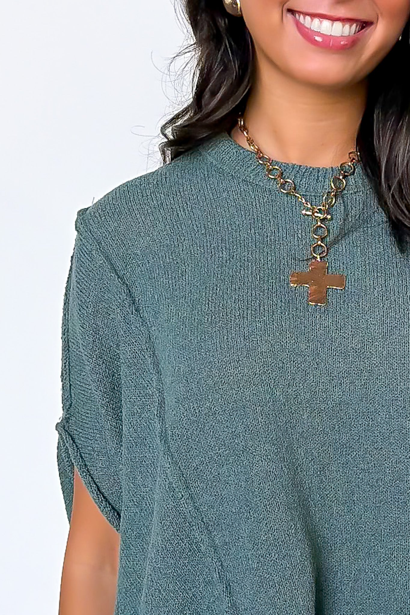 A close-up of a woman wearing a textured sage green knit top and a gold cross pendant necklace against a plain white background.